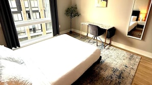 A hotel room with a large bed, a desk, a chair, a potted plant, and a large window offering a city view. - Luxury 1BD | 1 Bed | Northern Liberties (Philadelphia)