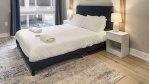 A neatly made bed with white linens and pillows, a bedside table with a lamp, and a large window with curtains. - Luxury 1BD | 2 Beds | Northern Liberties (Philadelphia)