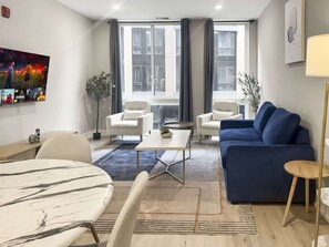 A modern living room with a blue sofa, white armchairs, a marble coffee table, and a large window with curtains.