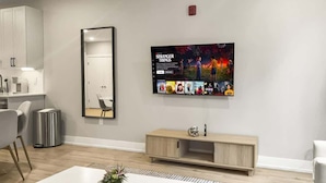 A modern hotel room with a white dining table, a small plant on it, a blue rug, a wooden cabinet, and a TV mounted on the wall displaying a movie poster. - Luxury 1BD | 2 Beds | Northern Liberties (Philadelphia)