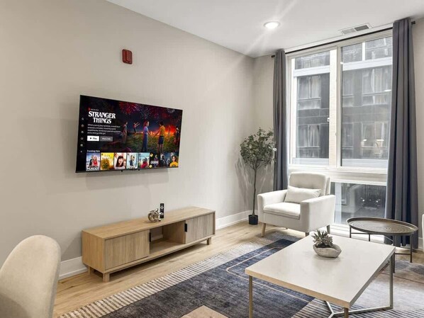 A modern living room with a flat-screen TV, a wooden console table, a white armchair, a round coffee table, and a large window with curtains.