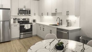 A modern kitchen with white cabinets, stainless steel appliances, a marble countertop, and a round dining table with a potted plant. - Luxury 1BD | 2 Beds | Northern Liberties (Philadelphia)