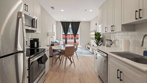 A modern kitchen with stainless steel appliances, a dining area with a wooden table and chairs, and a view of a red building through the window.