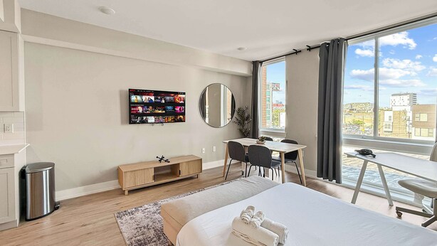 A modern hotel room with a large bed, a desk, a chair, a TV mounted on the wall, and a view of the city through the window. - Luxury Studio | 1 Bed | Northern Liberties (Philadelphia)