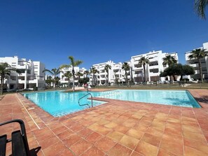 A swimming pool with tiled flooring, surrounded by white buildings and palm trees.