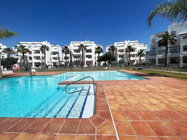 A swimming pool with clear blue water, surrounded by a tiled deck. There are palm trees and a row of white buildings in the background.