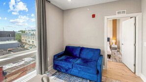 Een moderne hotelkamer met een blauwe veloursbank, uitzicht op de stad door grote ramen, en een houten vloer.