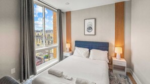 Een hotelkamer met een groot bed, twee nachtkastjes met lampen, uitzicht op gebouwen door het raam, en een ingelijste foto aan de muur.