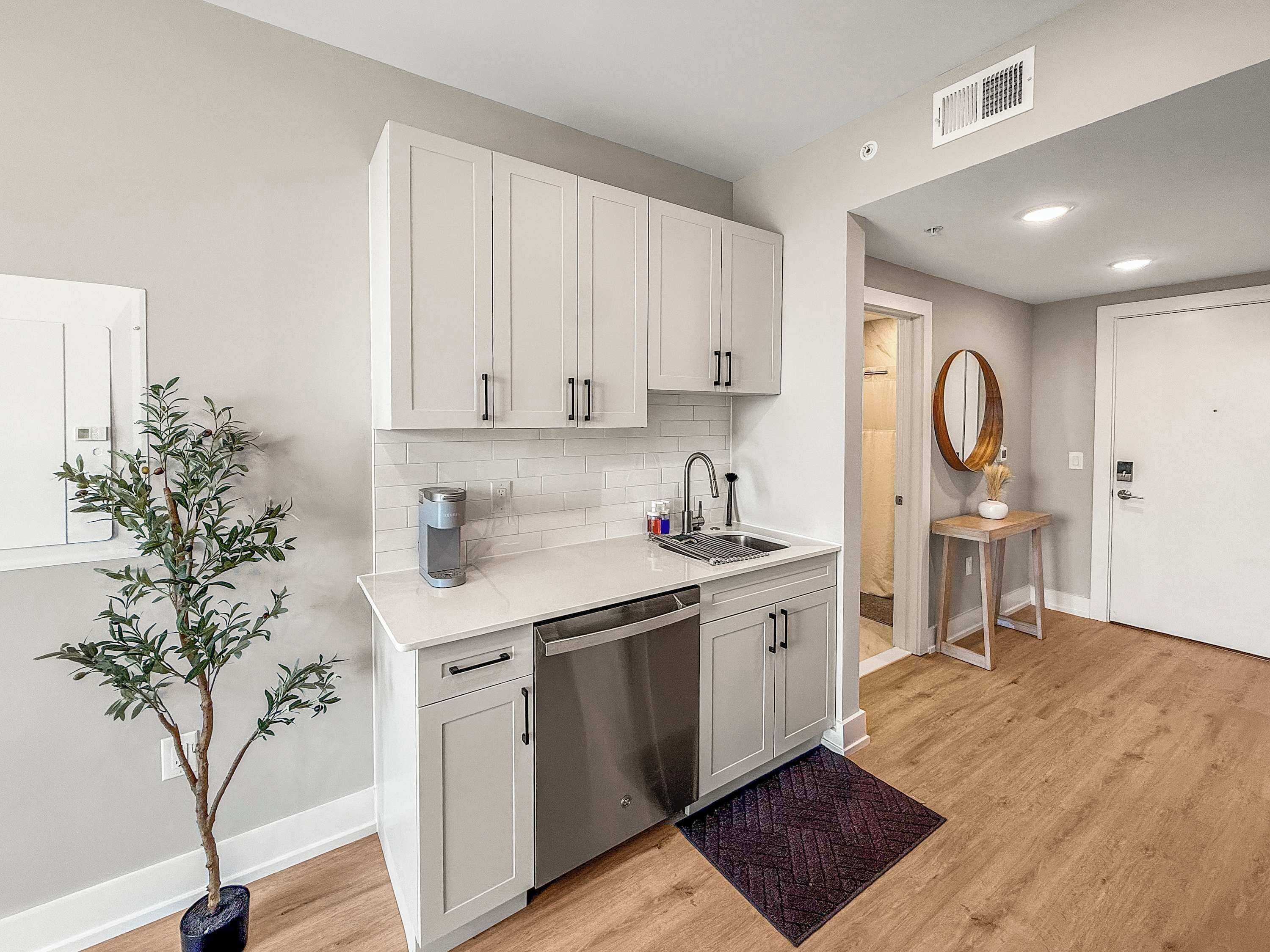 A modern kitchen with white cabinets, a built-in dishwasher, and a coffee maker. There is a potted plant and a wooden floor.