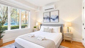 A modern bedroom with a large bed, bedside lamps, a window with a view, and an air conditioning unit.