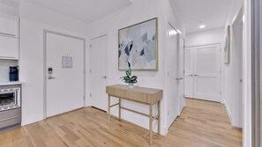 Käytävän lattia on parkettia, ovet ovat valkoiset, siellä on pieni puinen pöytä, jonka päällä on kasvi, ja seinällä on kehystetty abstrakti maalaus.