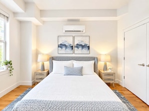 Un dormitorio con cama, dos mesitas de noche, una mesita auxiliar, una ventana con planta y una puerta.