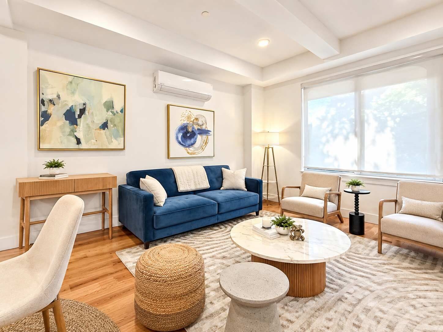 A modern living room with a blue sofa, round coffee table, and two armchairs. There are two paintings on the wall, a floor lamp, and a large window providing natural light.