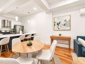 A modern kitchen with stainless steel appliances, a dining area with a round wooden table and chairs, and a living space with a blue sofa and a painting on the wall. - Modern Philly Haven | 1BD | Sleeps 4 | Northern Liberties (Philadelphia)