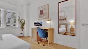 A hotel room with a bed, a desk with a computer, a chair, a potted plant, a large mirror, and artwork on the wall.