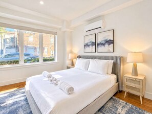 A hotel room with a large bed, two bedside tables with lamps, a window with a view of the street, and two abstract paintings on the wall. - Modern Philly Haven | 1BD | Sleeps 4 | Northern Liberties (Philadelphia)