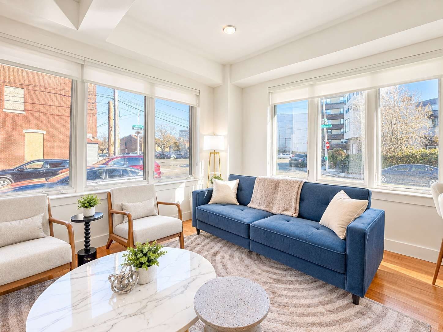 A modern living room with a blue sofa, two armchairs, a marble coffee table, and a view of the street through large windows.