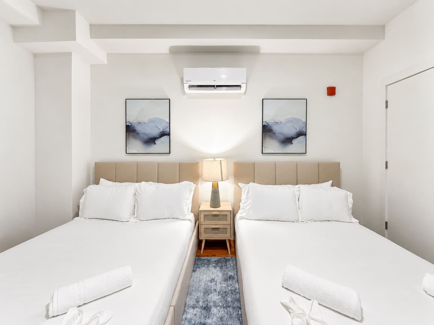 A hotel room with two beds, a nightstand, a lamp, and two abstract paintings on the wall.