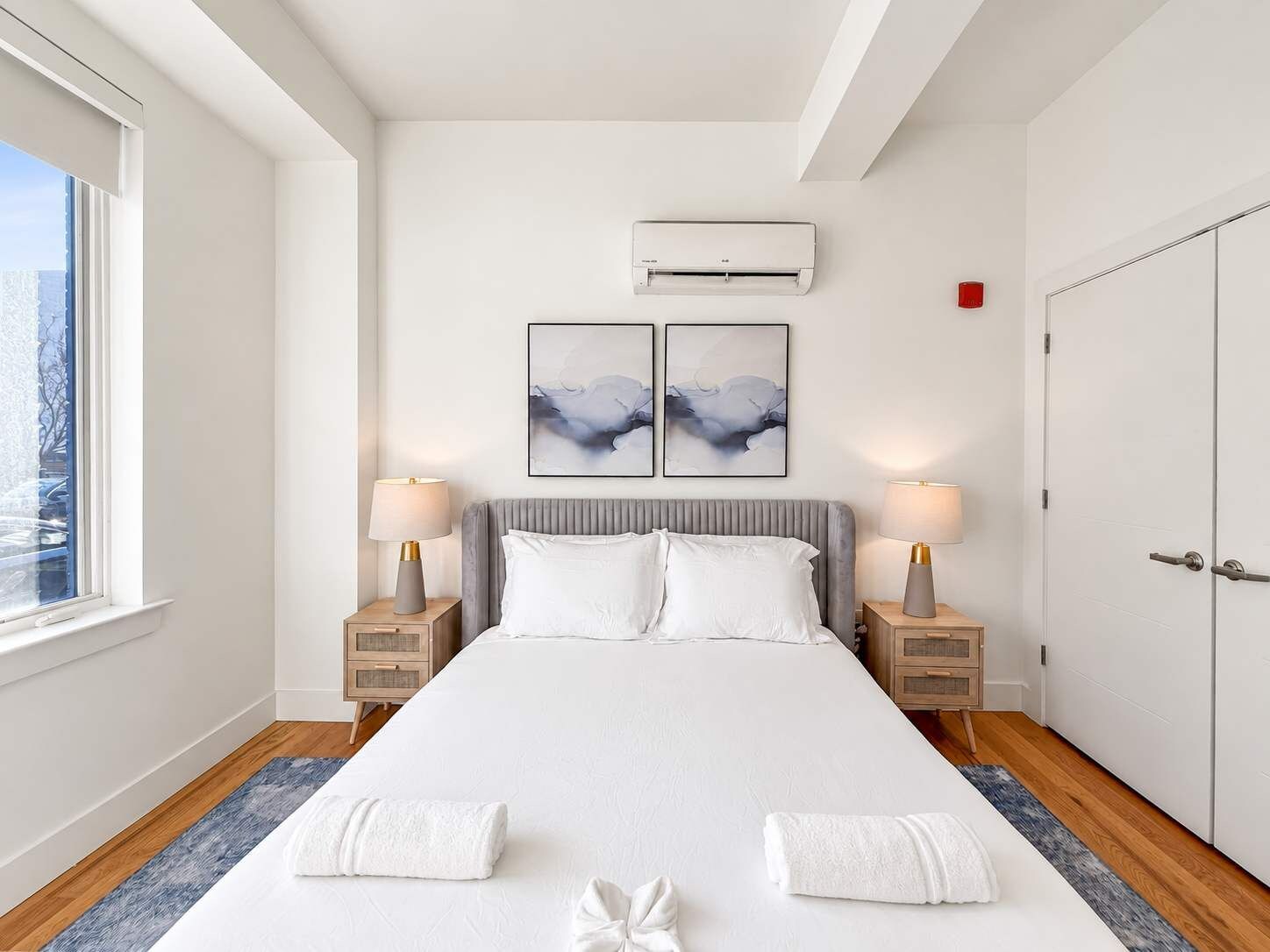 A hotel room with a large bed, two bedside tables with lamps, a window with a view, and an air conditioning unit on the wall.