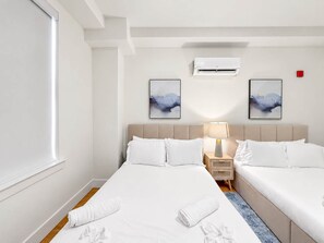 A hotel room with two beds, a window, an air conditioner, and two wall-mounted artworks. - Cozy Luxury 2BD | Sleeps 8 | Northern Liberties (Philadelphia)