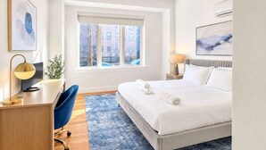 A modern hotel room with a large bed, a desk, a chair, a window with a view of buildings, and artwork on the walls.