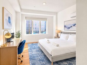 A modern hotel room with a large bed, a desk, a chair, a window with a view of buildings, and artwork on the walls.