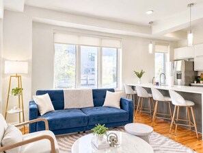 A modern living room with a blue sofa, a coffee table, and a dining area with bar stools. - Cozy Luxury 2BD | Sleeps 8 | Northern Liberties (Philadelphia)