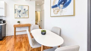 A modern dining area with a white table, grey chairs, and wooden flooring. There are two framed artworks on the walls.