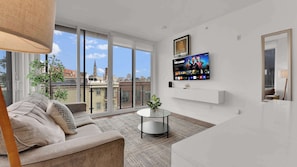 A modern living room with a sofa, a coffee table, a TV mounted on the wall, and large windows offering a city view. - Stylish 2BD | Sleeps 6 | Northern Liberties (Philadelphia)