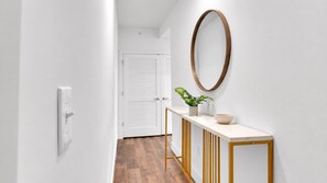 Un couloir avec une porte blanche, un plancher en bois, une console blanche sur laquelle sont posés un bol et une plante, et un miroir rond accroché au mur.