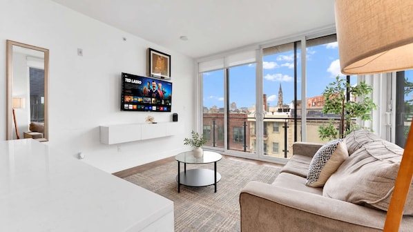 A modern living room with a flat-screen TV mounted on the wall, a round coffee table, a beige sofa, and a large window offering a city view.