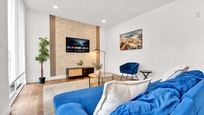 Un salon moderne avec un canapé bleu, un téléviseur à écran plat fixé au mur, une petite table ronde, une chaise bleue et un tableau.
