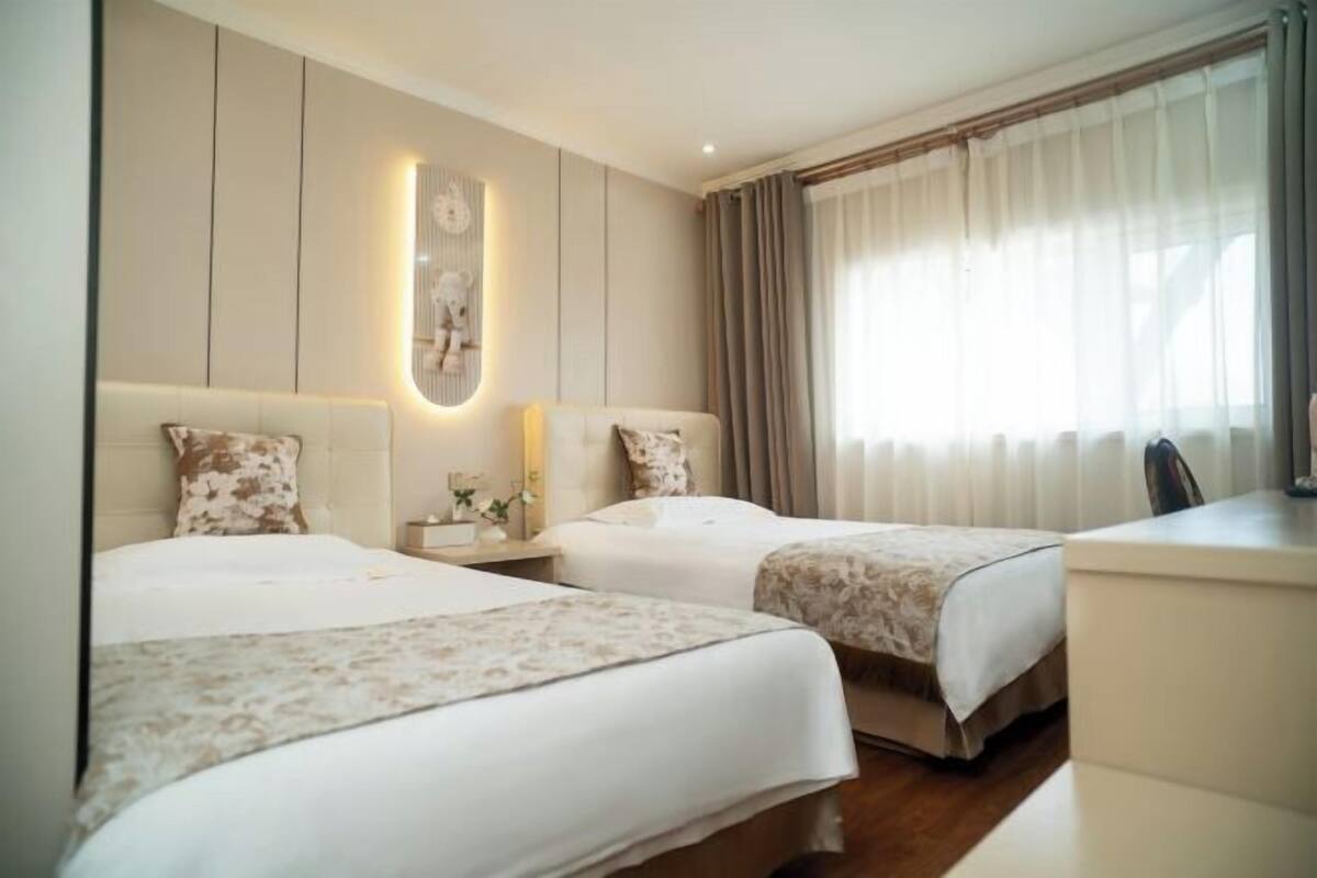 Camera d'albergo con due letti, una scrivania, una sedia, un comodino con lampada e un'ampia finestra con tende.