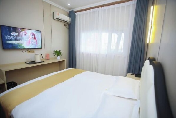 A hotel room with a bed, a TV mounted on the wall, a desk with a plant, and a window with curtains.