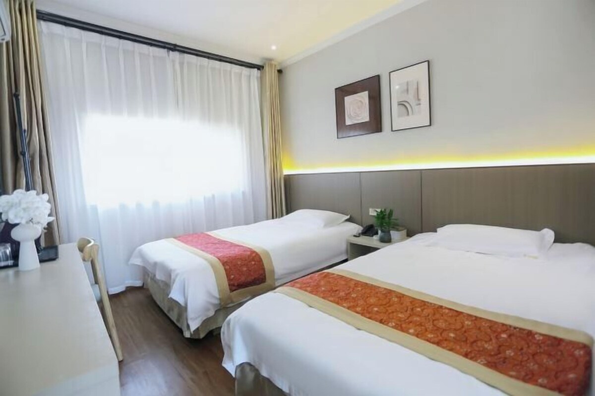 A hotel room with two beds, a desk with a chair, a window with curtains, and a bedside table with a plant.