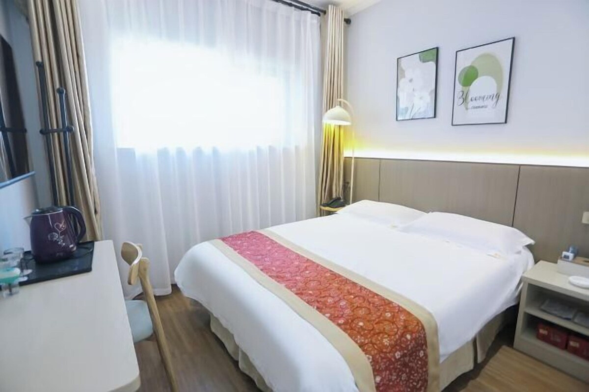 A hotel room with a bed, a desk with a kettle, a chair, a lamp, and two framed pictures on the wall.