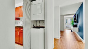 Een moderne keuken met een ingebouwde wasmachine en droger, houten kastjes en uitzicht op de stad door een open deur.