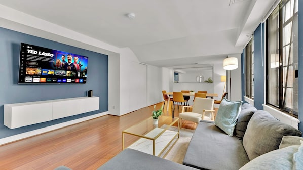 A modern living room with a flat-screen TV mounted on the wall, a sofa, a coffee table, and a dining area with chairs and a table.
