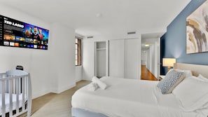 A hotel room with a large flat-screen TV mounted on the wall, a bed with white linens, a bedside table, and a crib.