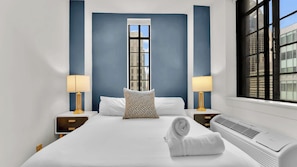 A hotel room with a large bed, two bedside tables with lamps, a window with a view of buildings, and a wall-mounted air conditioner.