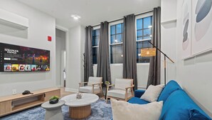 A modern living room with a blue sofa, white armchairs, a coffee table, and a TV displaying 'Stranger Things'. - Modern 3-Bedroom Oasis in Center City (Philadelphia)