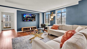 A living room with a sofa, a coffee table, a TV mounted on the wall, and a window with blinds.