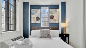 A hotel room with a large bed, a window, a wall-mounted air conditioner, and two abstract paintings.