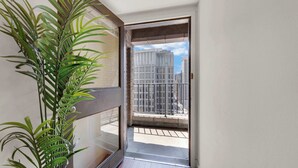 Un balcón con vistas a edificios altos, una planta en maceta y una puerta que da al interior.