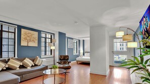 Ein modernes Wohnzimmer mit Sofa, Sessel und Couchtisch. Zwei Fenster mit Jalousien, ein Bild an der Wand und eine Tür zu einem anderen Raum.