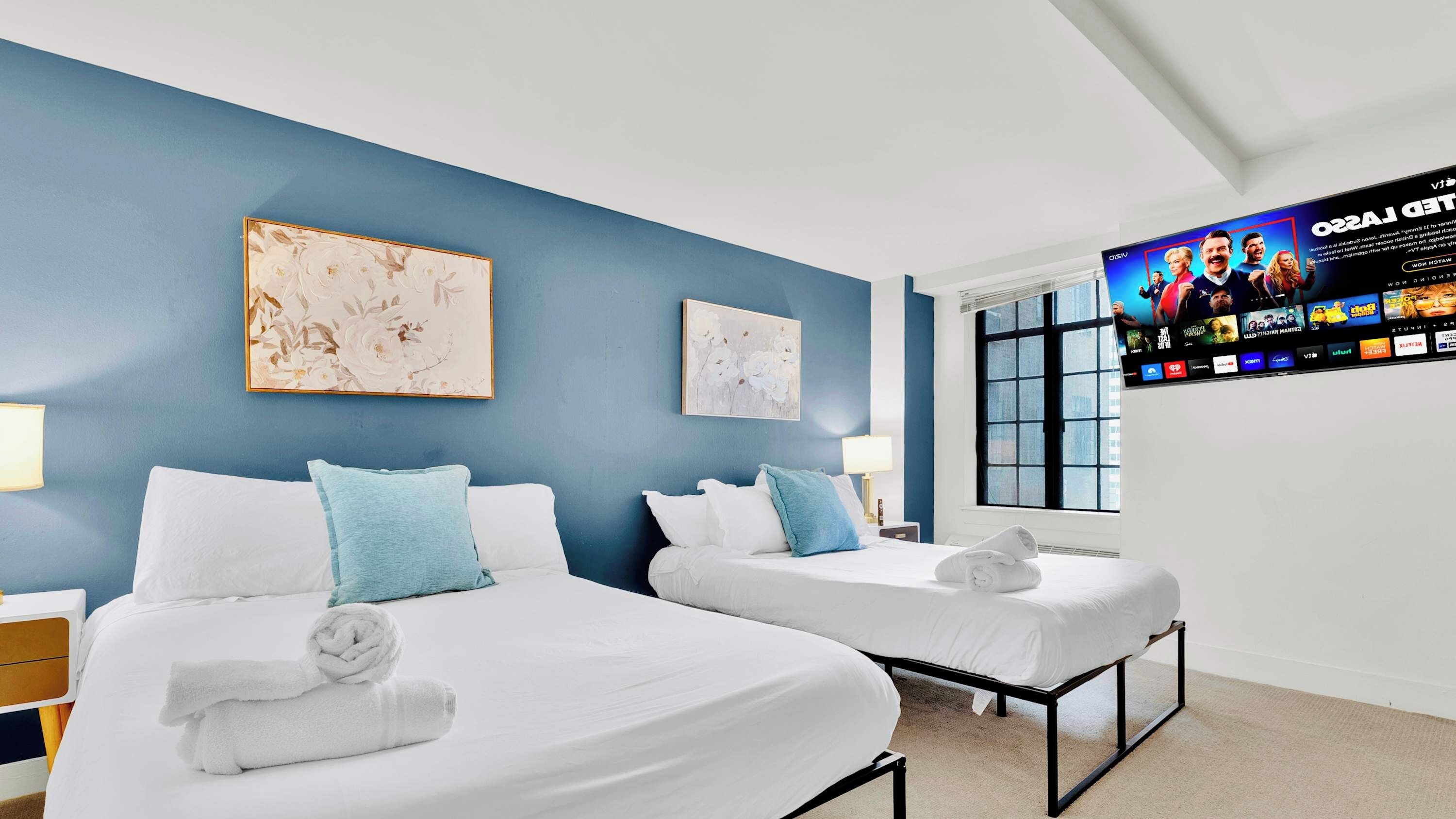 Une chambre d’hôtel avec deux lits, un téléviseur fixé au mur et des œuvres d’art accrochées aux murs.