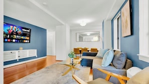 A modern living room with a flat-screen TV, a dining area with a table and chairs, and a sofa with cushions.
