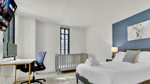 Quarto de hotel com cama, escrivaninha, cadeira, radiador, televisão e janela.