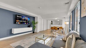 A modern living room with a flat-screen TV, a dining area, and a kitchen in the background.
