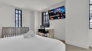 A hotel room with a bed, a desk with a computer, a TV on the wall, and large windows.